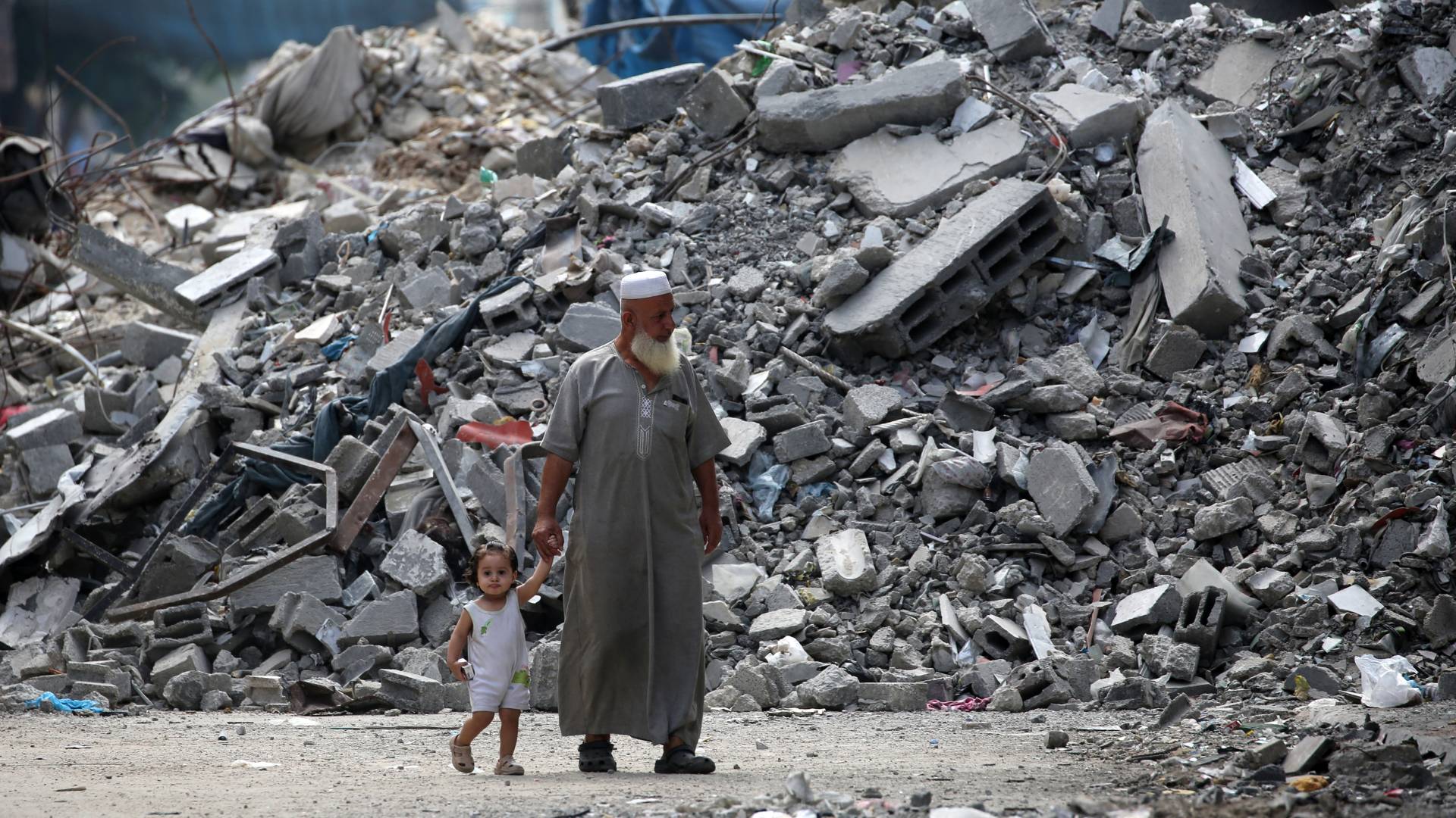 رجل مسن يحمل طفلًا بيده وهو يسير بجوار مبنى دمره قصف الاحتلال في مخيم البريج للاجئين بوسط قطاع غزة
