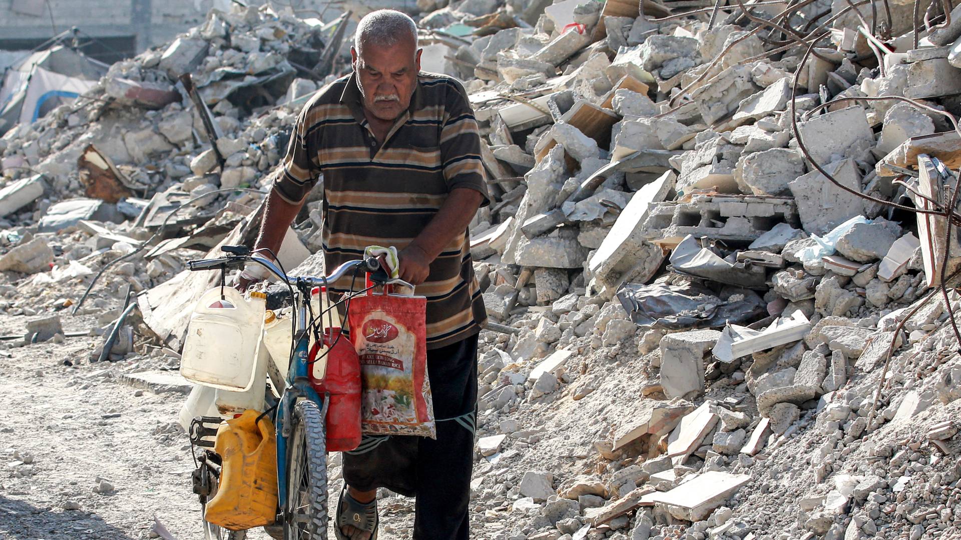 رجل يدفع دراجة محملة بحاويات لملء المياه أمام أنقاض مبنى منهار في خان يونس بجنوب قطاع غزة