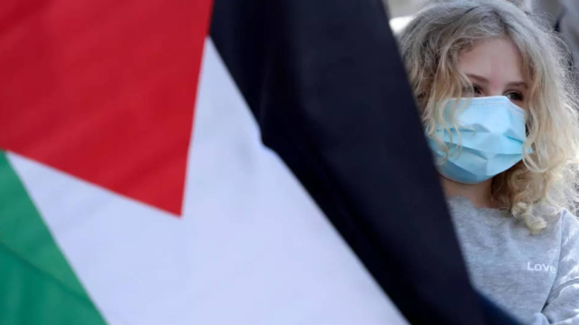 فتاة فلسطينية تضع كمامة واقية من فيروس وكورونا وترفع العلم الفلسطيني