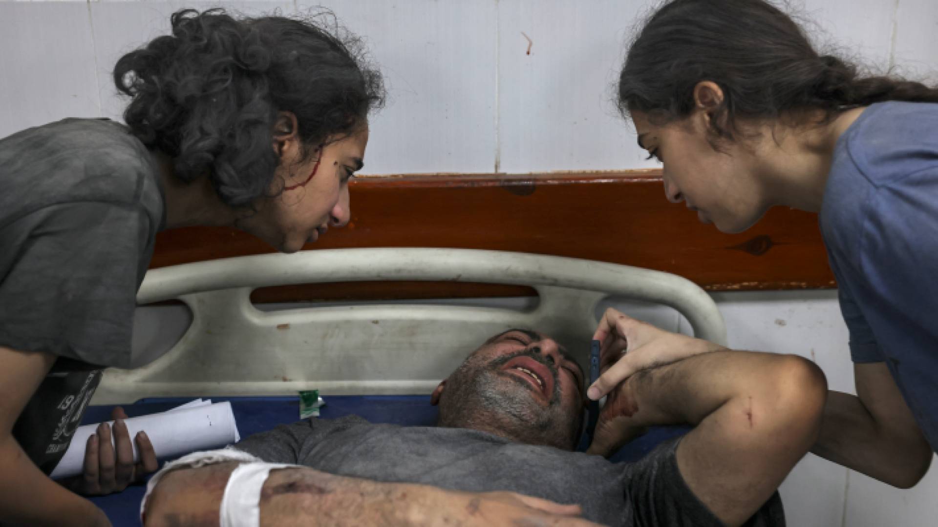 طفلتان أمام والدهما الذي يتلقى العلاج عقب إصابته بقصف الاحتلال الإسرائيلي