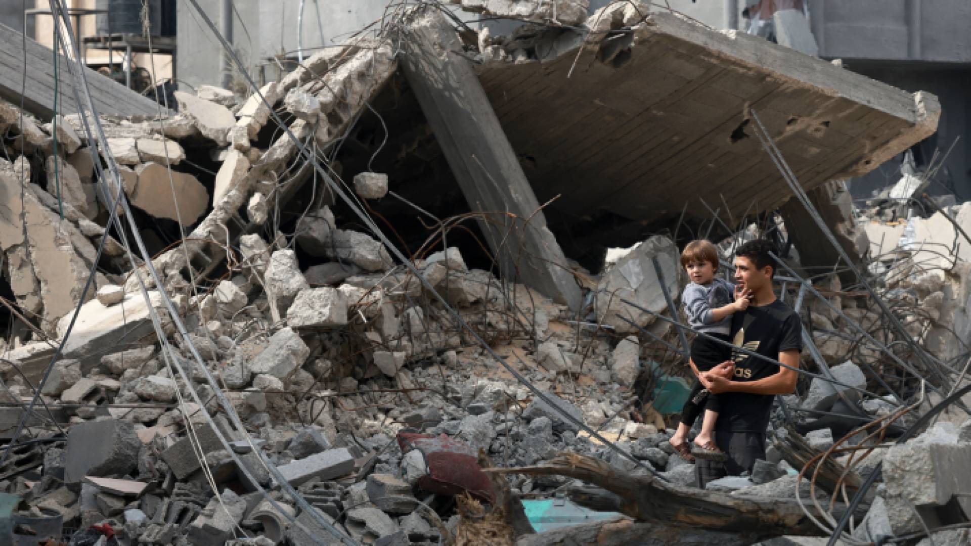 فتى فلسطيني يحمل طفل بالقرب من أحد المنازل التي دمرت بقصف الاحتلال في غزة