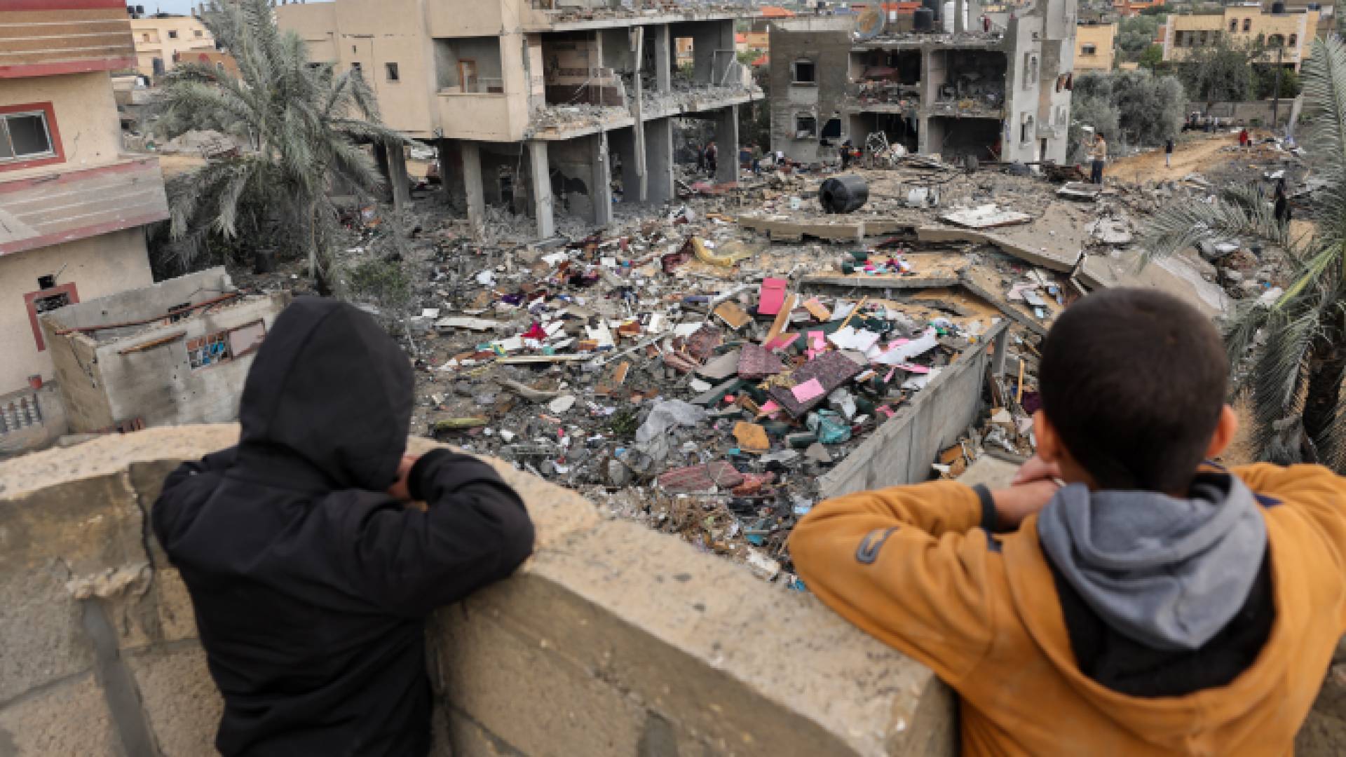 طفلان فلسطينيان ينظرون بينما يقف الناس على أنقاض مبنى دمر خلال الغارات الإسرائيلية