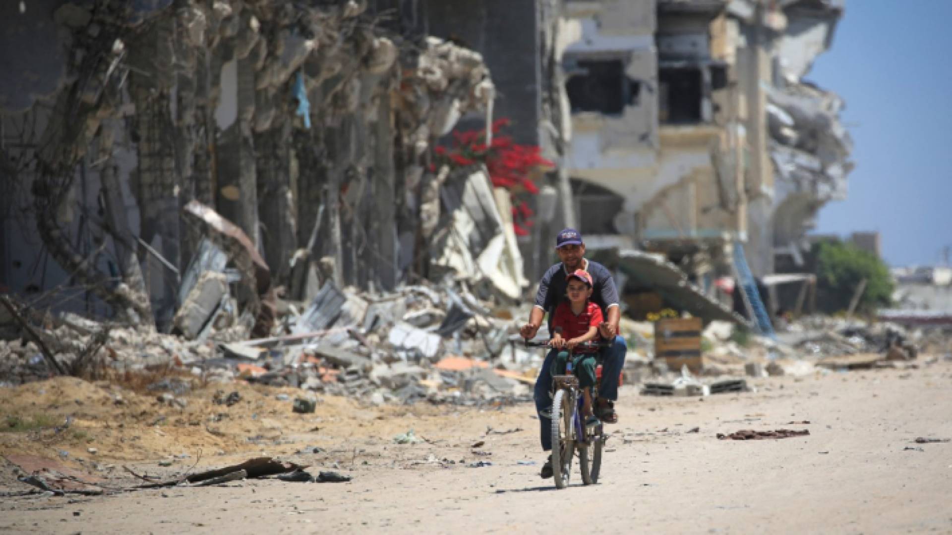 رجل مع طفل يجلس على عارضة دراجة، يمر عبر المباني المدمرة في خان يونس