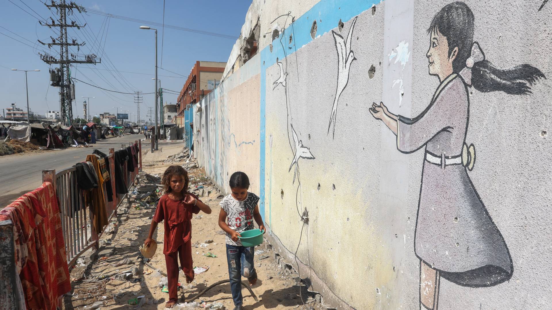 فتيات فلسطينيات يسيرن أمام جدارية في مخيم نازحين مؤقت أقيم على جانب الطريق في دير البلح وسط قطاع غزة